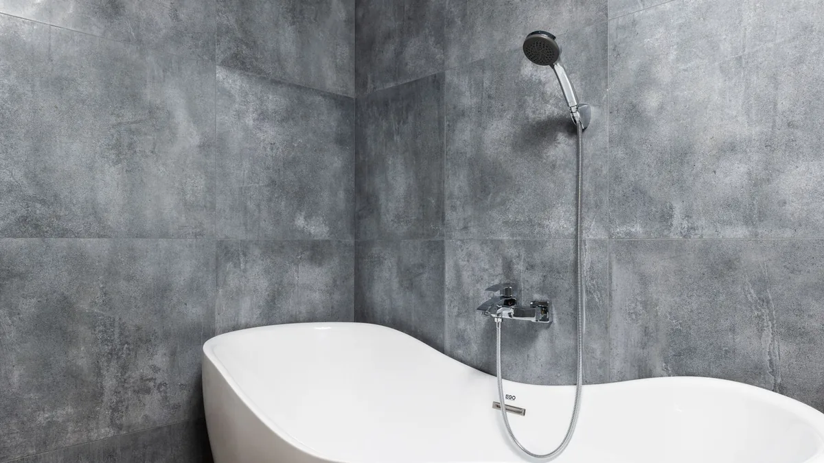 Bañera exenta blanca junto a una pared de baldosas grises, con grifo mezclador y ducha de mano montados en la pared.