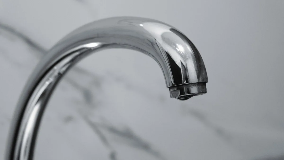 Primer plano del caño curvado de un grifo cromado, con una gota de agua en la boquilla, sobre un fondo de mármol.