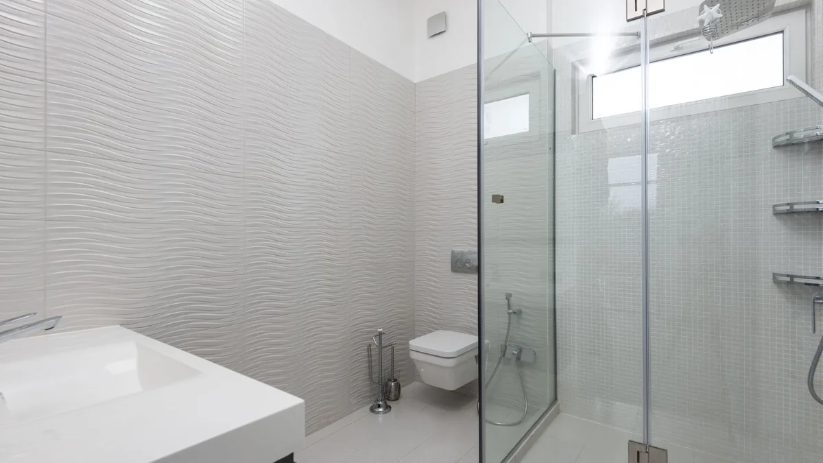 Cabina de ducha con puertas de cristal abatibles, en un baño con pared alicatada de textura ondulada e inodoro suspendido al fondo.
