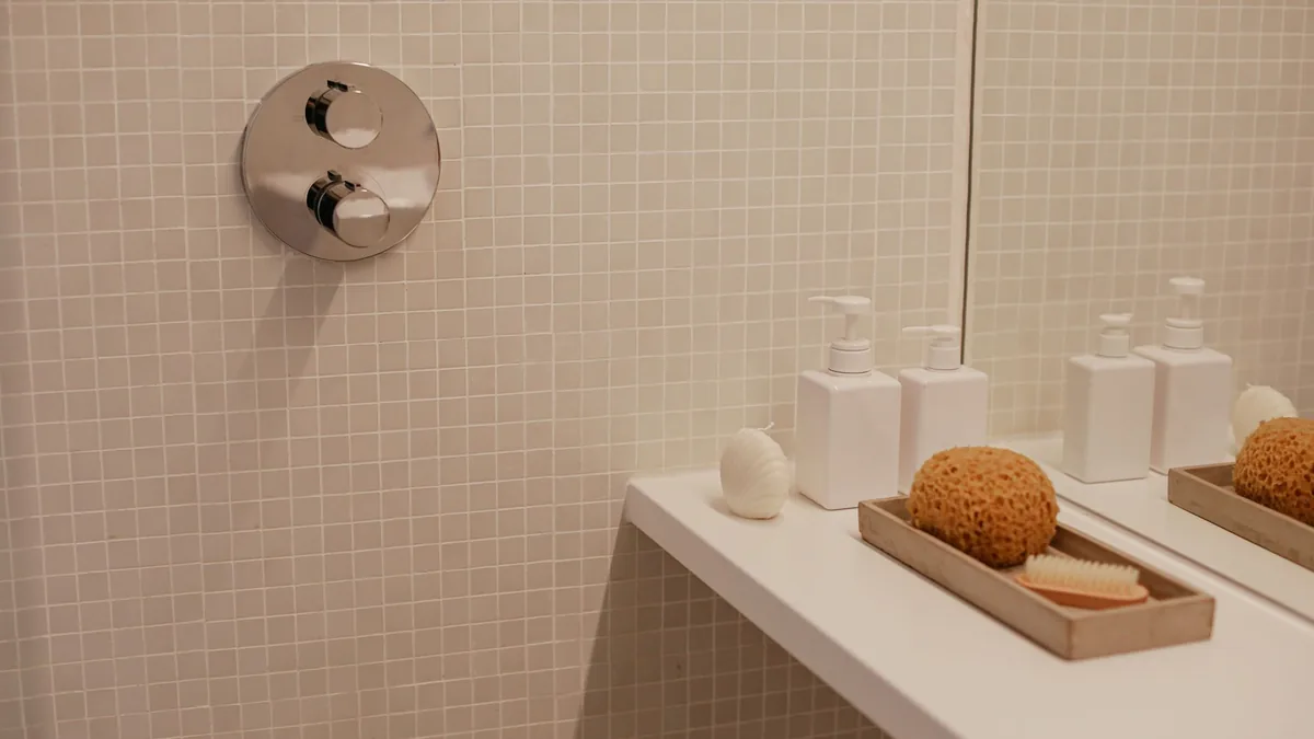 Mando de ducha termostático empotrado en una pared alicatada, junto a dosificadores de jabón y una esponja natural sobre la encimera del lavabo.