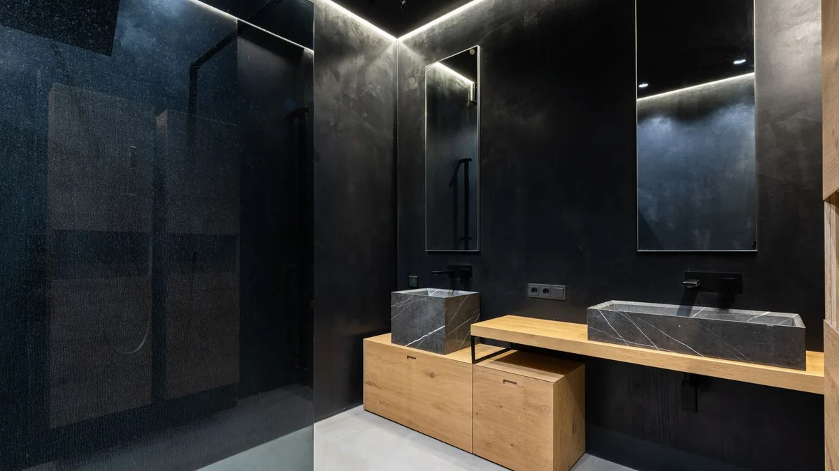 Mueble de baño de madera con dos lavabos de piedra oscura sobre la encimera, grifos negros y espejos con luz integrada.