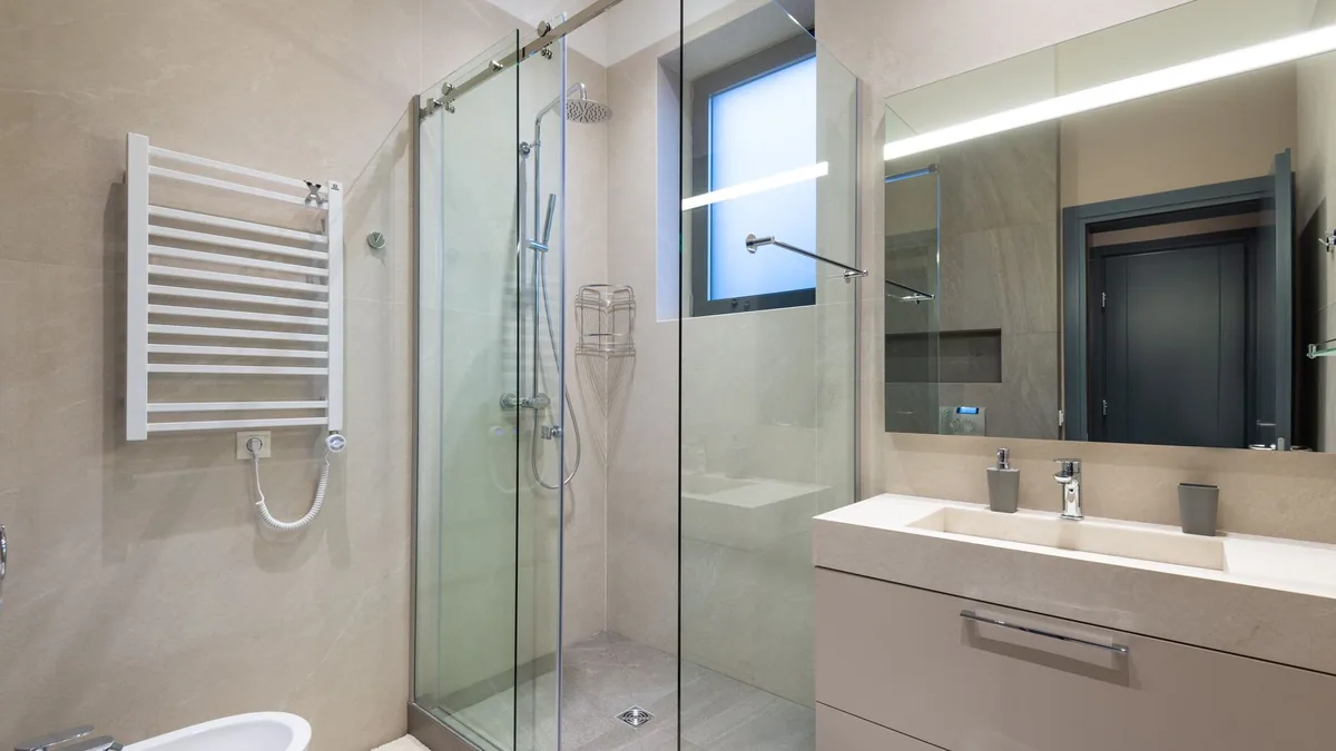 Baño moderno con radiador toallero blanco a la izquierda, mampara de ducha corredera de cristal en el centro y lavabo con espejo iluminado a la derecha.