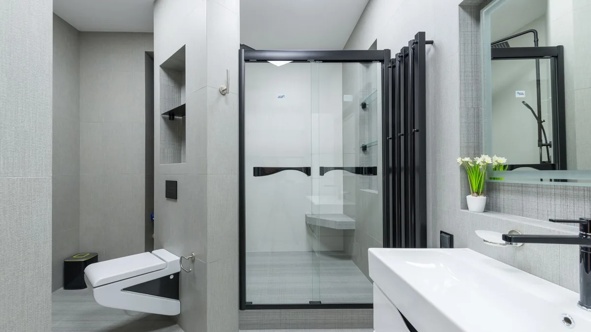 Inodoro suspendido en un baño con ducha de cristal al fondo y lavabo con grifo negro a la derecha.